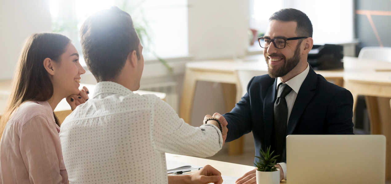 Un homme en costume sourit et serre la main d’un autre homme de l’autre côté d’un bureau. Une femme est assise à côté d’eux, souriante.