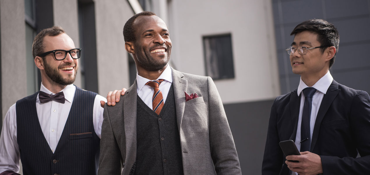 Trois hommes de divers horizons en tenue formelle, riant et discutant en marchant.
